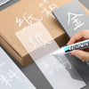 A hand holds a white permanent paint marker pen next to various transparent and opaque markers in a cardboard box.