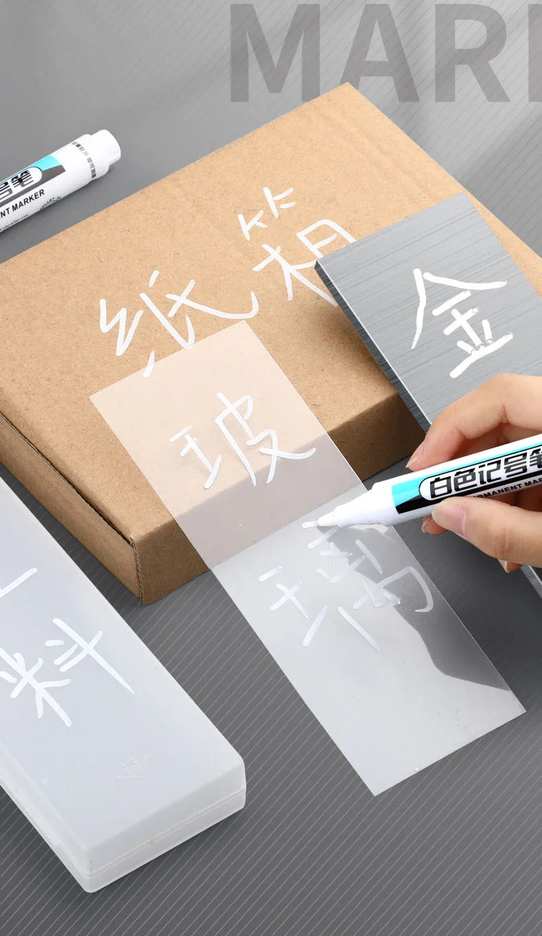 A hand holds a white permanent paint marker pen next to various transparent and opaque markers in a cardboard box.