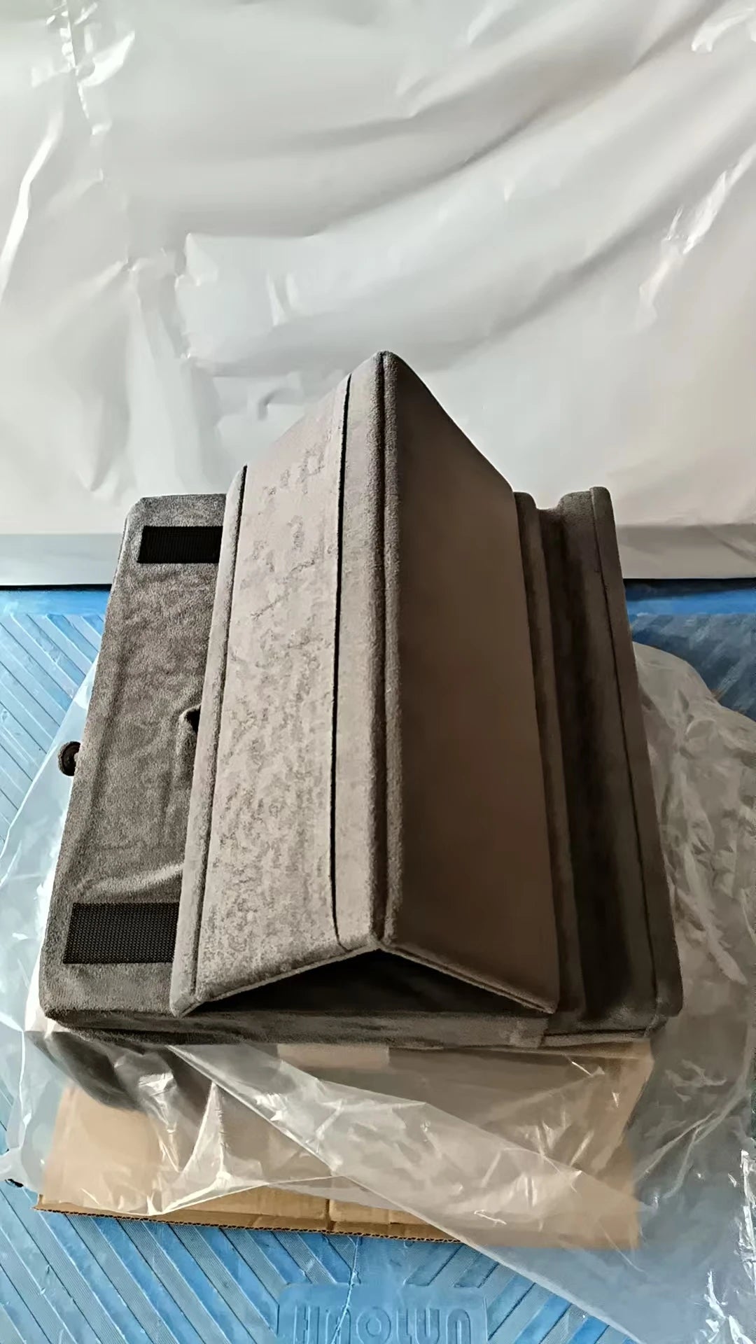 A gray foldable tablet pillow stand with a pocket is shown resting on a cardboard box inside clear plastic packaging.