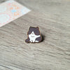 Un pequeño broche de esmalte en forma de un gato sonriente y tierno con marcas marrones y blancas, sobre una superficie de ma