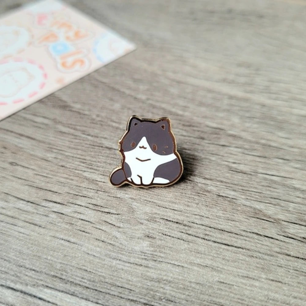 Un pequeño broche de esmalte en forma de un gato sonriente y tierno con marcas marrones y blancas, sobre una superficie de ma