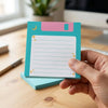 A hand holds a teal sticky note with a pink header and white lined space, featuring a cosmic moon design.