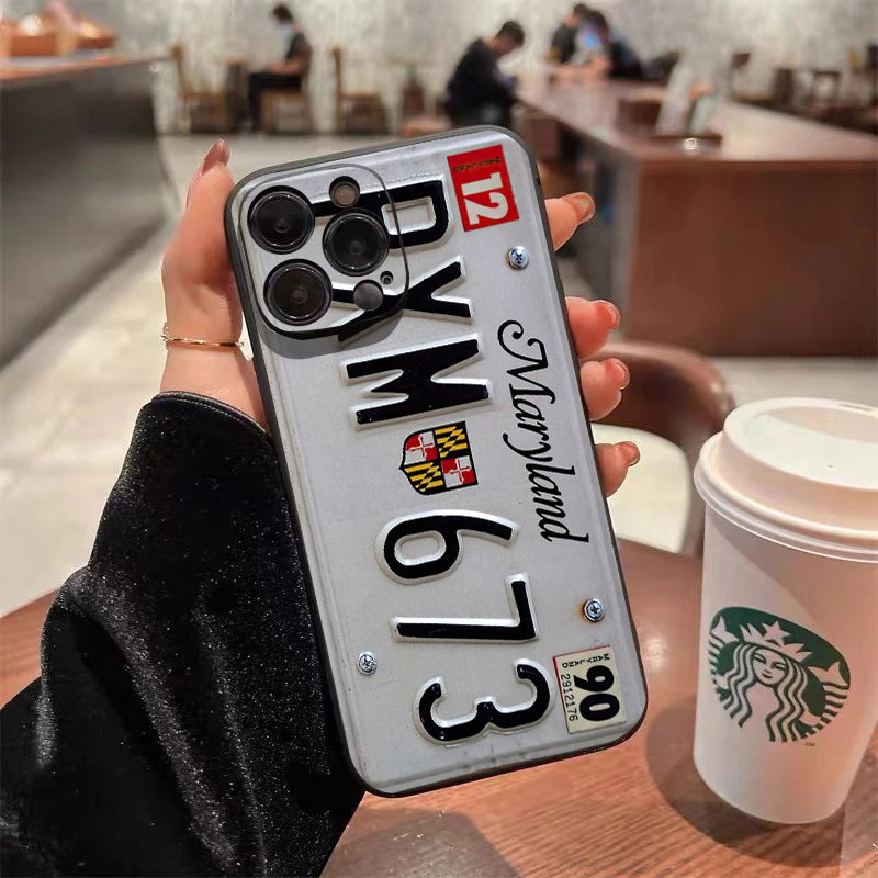 A hand holds a silver vintage license plate phone case for an iPhone with Maryland text and a green Starbucks cup in the back