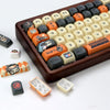 A close-up view of a brown mechanical keyboard featuring orange and black PBT keycaps, along with various custom keycaps and 