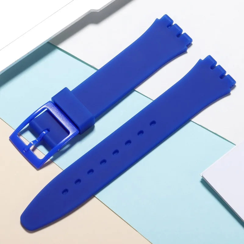 A blue silicone watch strap with a buckle and replacement tools is displayed on a light background.