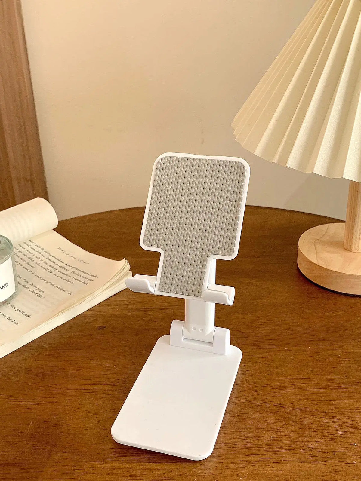 A white adjustable foldable phone and tablet stand resting on a wooden table next to an open book.