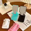 A pink adjustable foldable phone and tablet stand is shown on a wooden table next to a notebook.