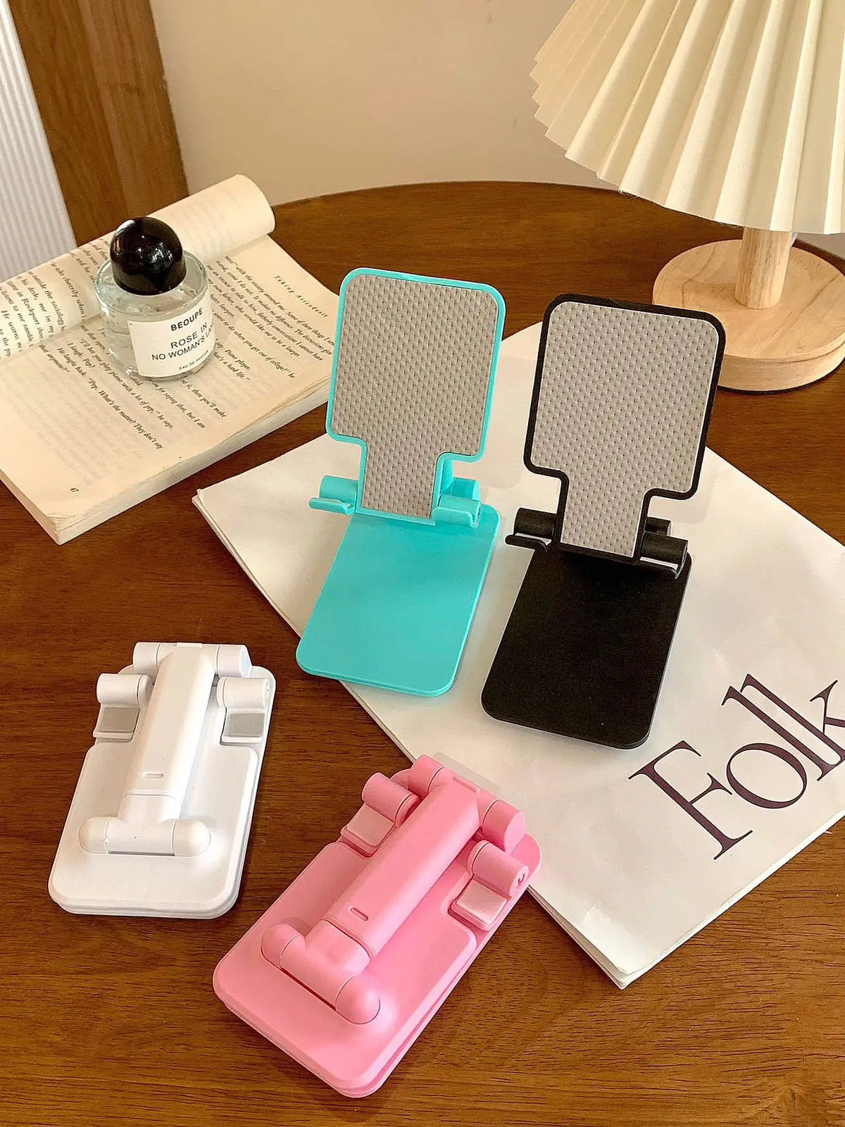 A pink adjustable foldable phone and tablet stand is shown on a wooden table next to a notebook.