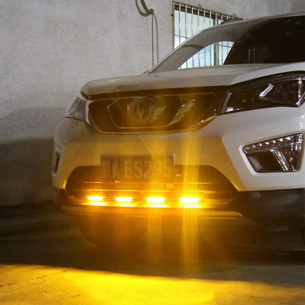 A white car's headlights are brightly illuminated, casting a strong yellow glow on the ground.