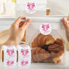 Eine Hand legt rosa Dankeschön-Sticker auf eine durchsichtige Cellophan-Tüte voller Croissants.