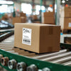 A brown shipping box labeled USPS Priority Mail resting on a conveyor belt with rolls of thermal shipping labels.