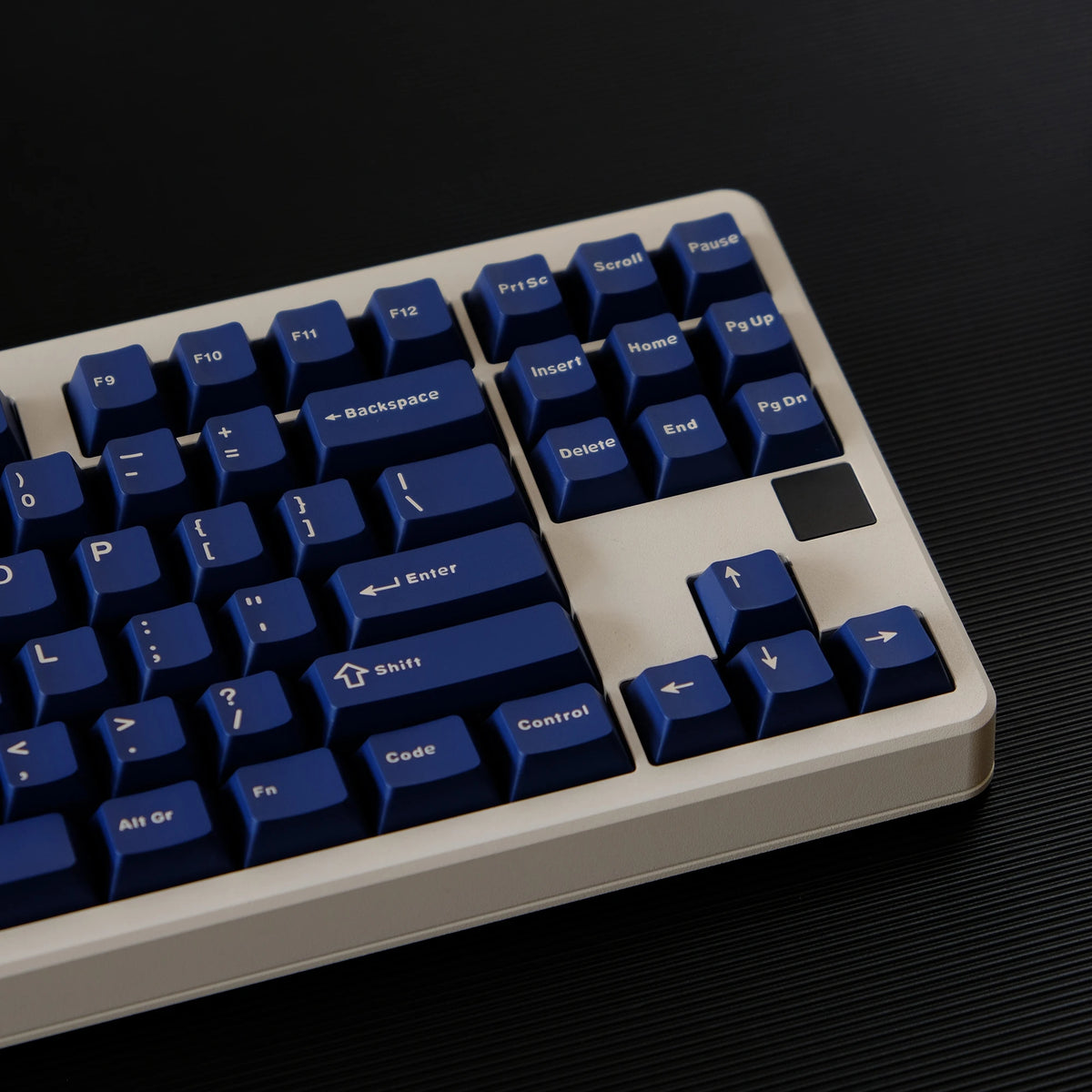 A close-up view of a light beige mechanical keyboard with dark blue PBT keycaps and a black keycap, shown on a dark surface.