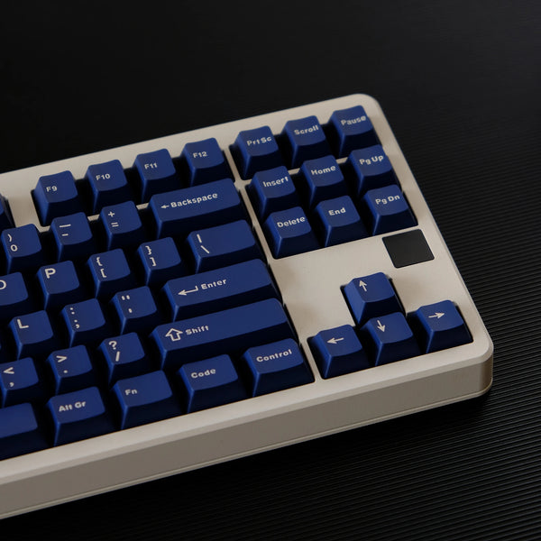 A close-up view of a light beige mechanical keyboard with dark blue PBT keycaps and a black keycap, shown on a dark surface.