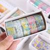 A hand holds an open box of decorative transparent washi tape rolls featuring floral patterns and pastel colors.