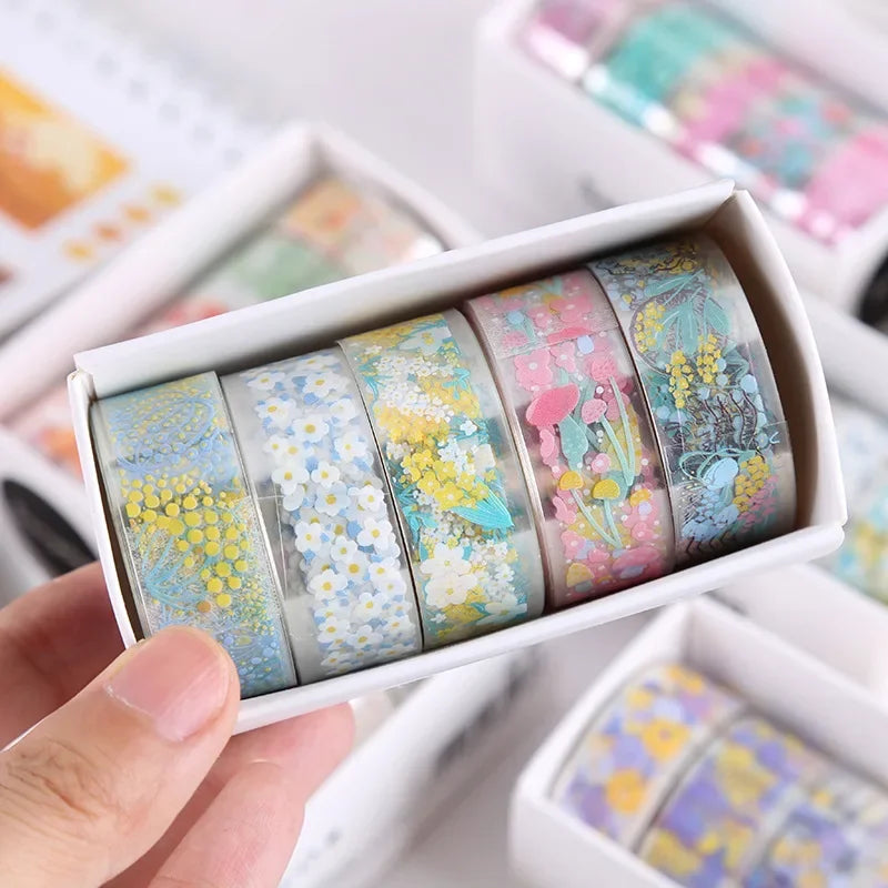 A hand holds an open box of decorative transparent washi tape rolls featuring floral patterns and pastel colors.