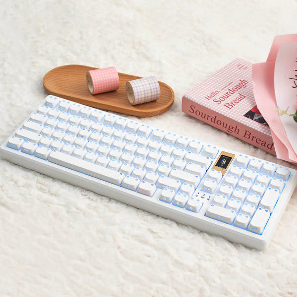 Un teclado mecánico blanco con teclas rosas, junto a pan decorativo rosa y teclas de tela enrolladas.