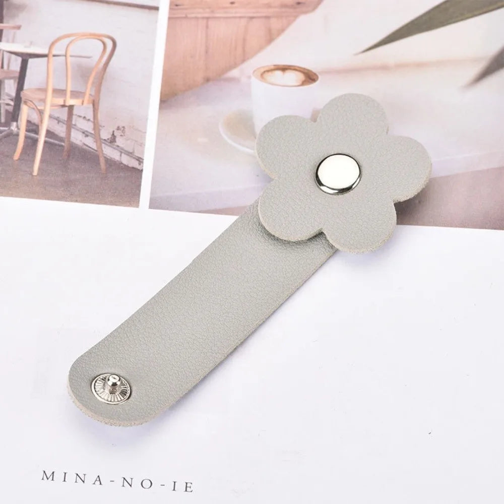 A gray leather flower cable organizer with a silver stud, shown next to a blurred background of wooden furniture.