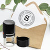 A black self-inking stamp with a circular logo and text is placed on a brown envelope next to a stamp pad.