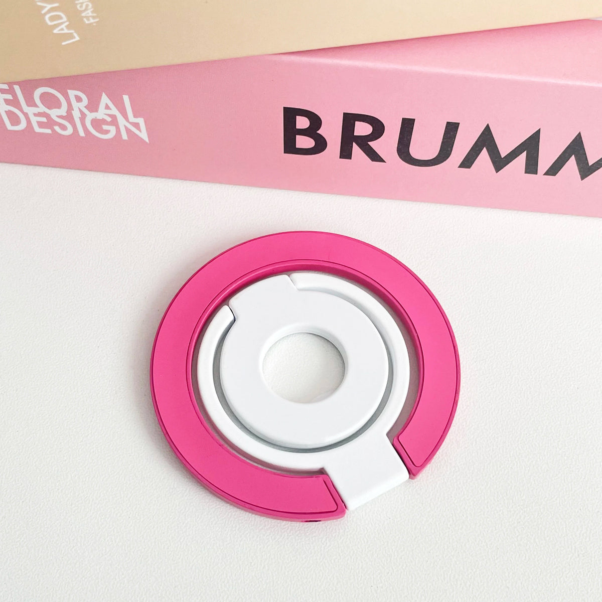 A pink magnetic phone grip and stand with a foldable ring resting on a white surface.