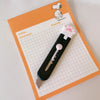 A black mini cutter with a white handle and pink paw tip rests on an orange sheet of graph paper.