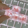 Small translucent pink resin keycaps shaped like botanical elements resting on a light-colored mechanical keyboard.