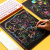 A hand draws colorful, whimsical characters on a black art paper sheet using stylus sticks.