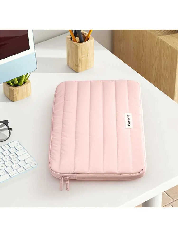 A pink padded laptop sleeve resting on a white desk next to a tablet and wooden containers.