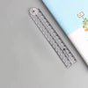 A transparent folding ruler set with a circular marker is shown on a light gray surface next to a striped notebook.