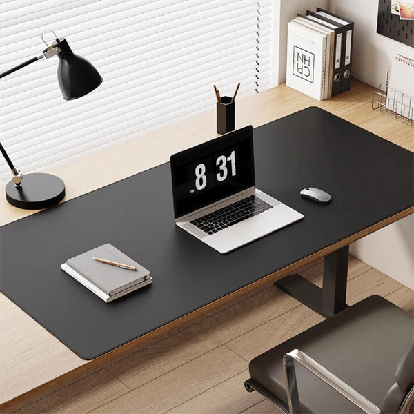 A black laptop is placed on a dark desk mat in an office setting with a lamp and books visible.