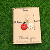 A thank you card with dimensions and a ladybug and daisy keychain set displayed on green grass.