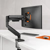 A black fully adjustable monitor arm is mounted on a desk, positioned next to a computer monitor.