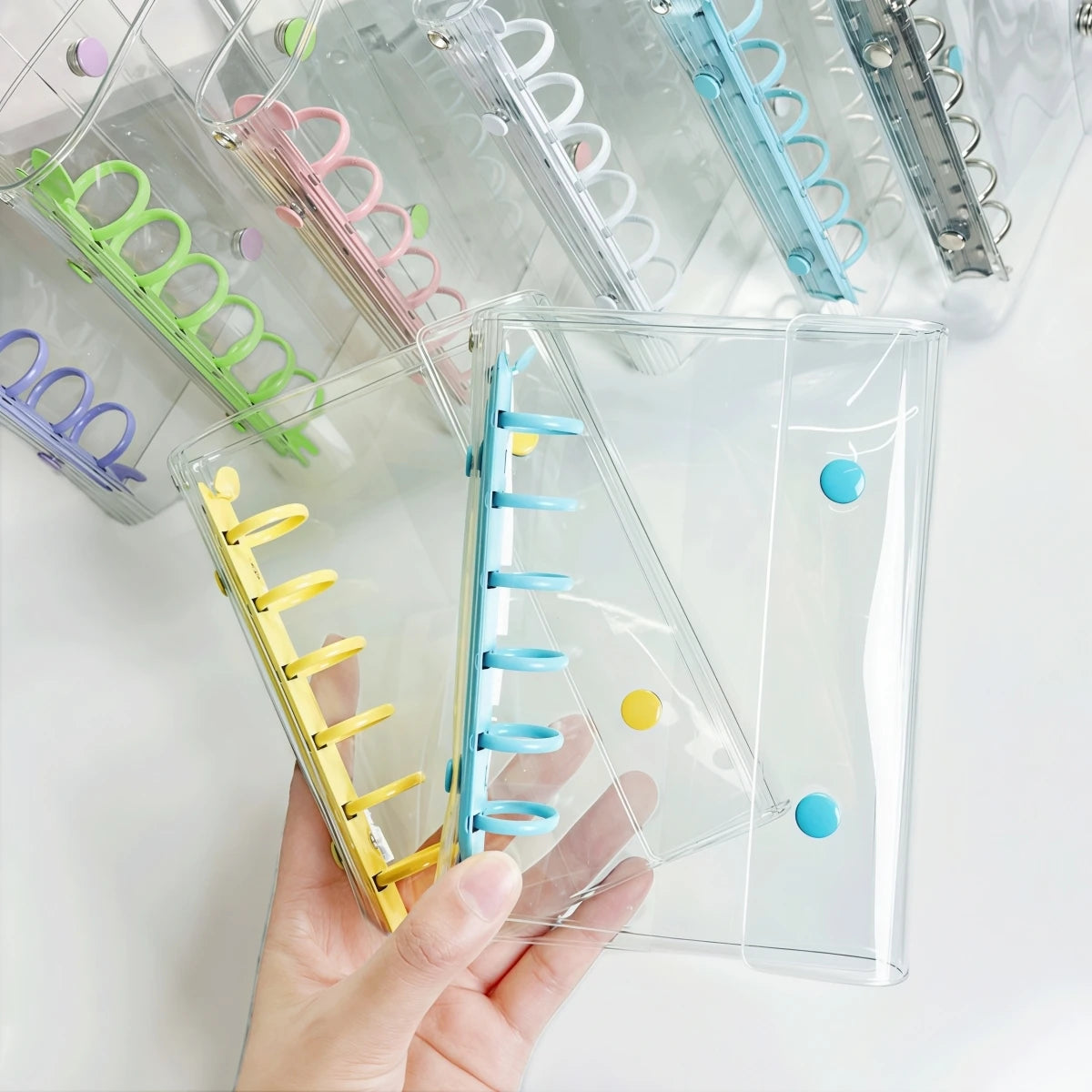 A hand holds a clear double-button colorful binder for stickers and journaling with various colored plastic rings.