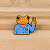 An enamel pin featuring a sarcastic frog wearing a blue shirt with a red star emblem and making a peace sign.