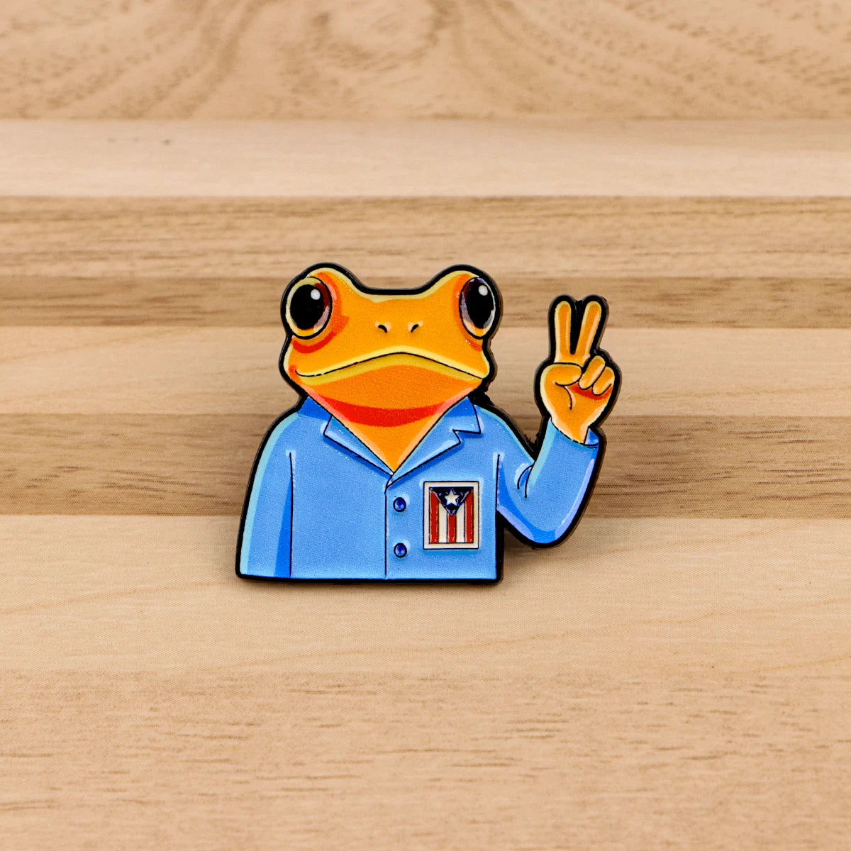 An enamel pin featuring a sarcastic frog wearing a blue shirt with a red star emblem and making a peace sign.
