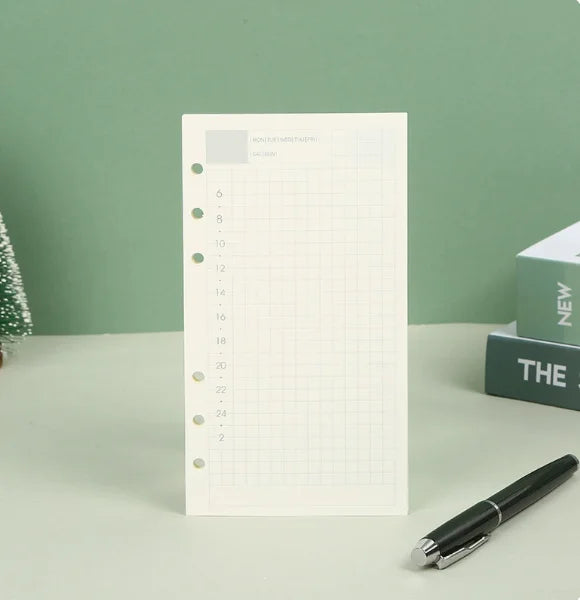 Un cuaderno de hojas sueltas blanco con cuadrícula y líneas numeradas junto a un bolígrafo negro sobre una superficie verde c
