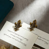 A set of two decorative iris-shaped gold metal binder clips resting on a white card against a gray background.