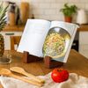 A vintage wooden cookbook holder stands on a wooden table next to a cookbook, a tomato, and wooden spoons.