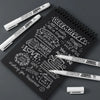 Three white permanent paint markers resting on a black spiral notebook with handwritten December messages.