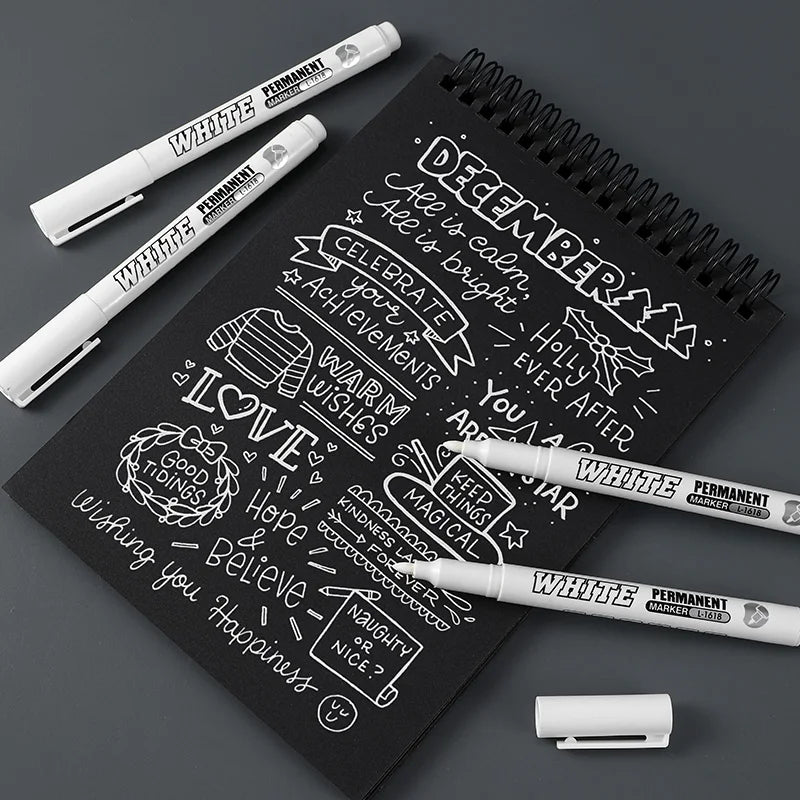 Three white permanent paint markers resting on a black spiral notebook with handwritten December messages.