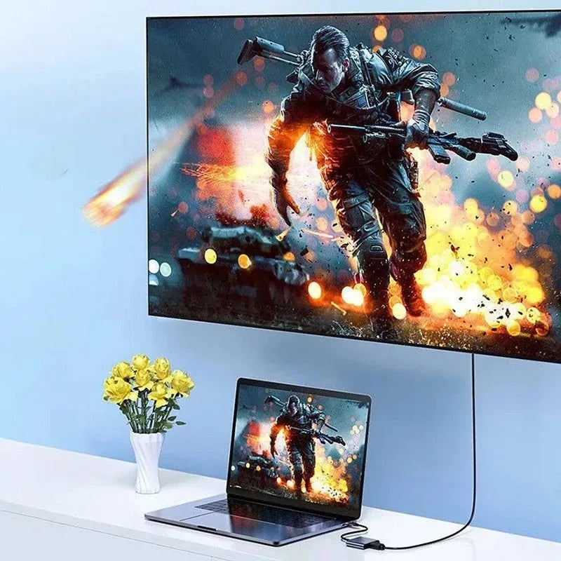 A laptop is placed in front of a large monitor displaying an action scene, with a 3-in-1 USB-C to HDMI docking adapter visibl