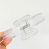 A hand holds a clear plastic book page clip with a silver spring mechanism.