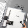 Un carnet à feuilles transparentes avec une reliure spirale noire, à côté de lunettes et d'un stylo.
