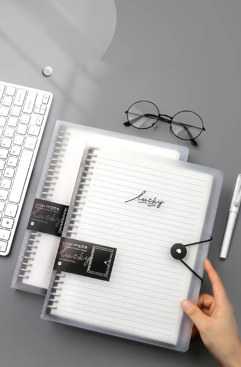 Un carnet à feuilles transparentes avec une reliure spirale noire, à côté de lunettes et d'un stylo.