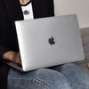 Eine Person hält einen silbernen Laptop mit Schutzhülle und spiegelndem Finish vor einem hellen Hintergrund.
