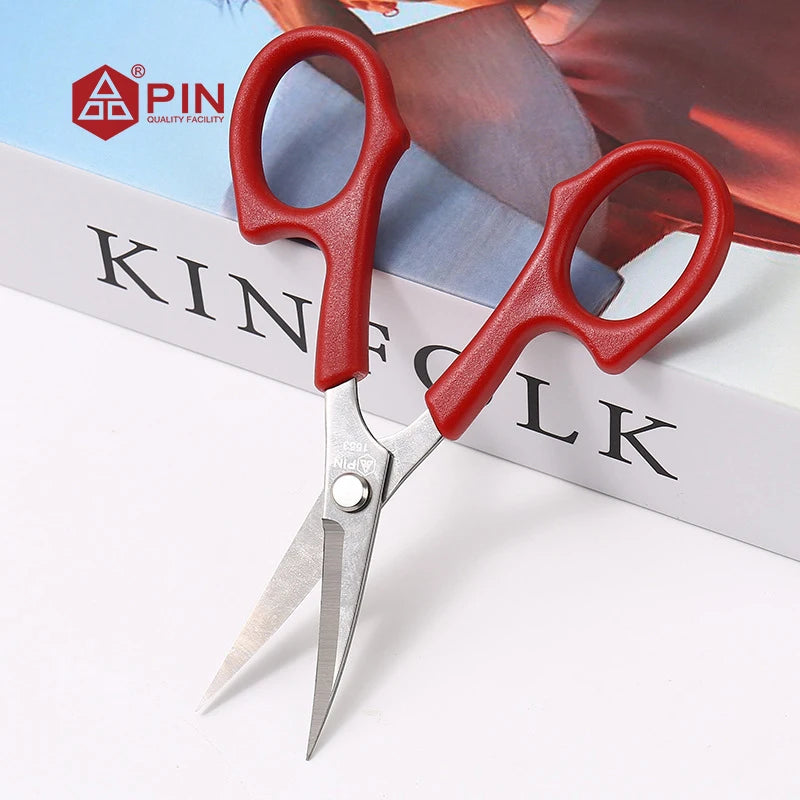 A pair of red embroidery scissors with silver blades resting on a white background next to a book.