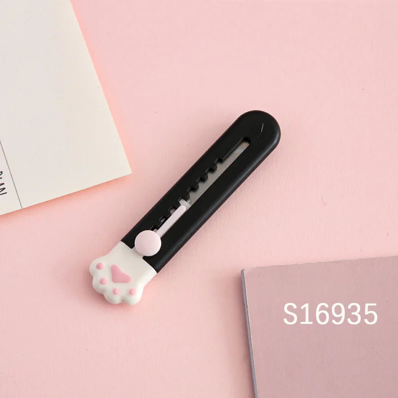 A black portable pet claw tool knife with a white paw print decoration rests on a pink surface next to a pink notebook.