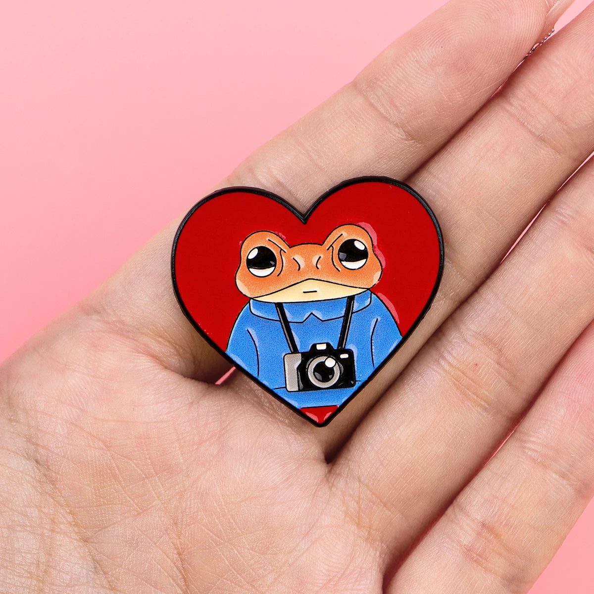 An enamel pin featuring a cartoon frog wearing a blue shirt and carrying a camera, set against a red heart background.
