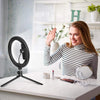 A smiling woman demonstrates a 12 inch LED ring light with a tripod stand and remote control on a white table.