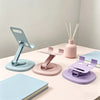 A pink and blue 360° rotating foldable desktop phone stand is shown on a white table next to a notebook and chopsticks.