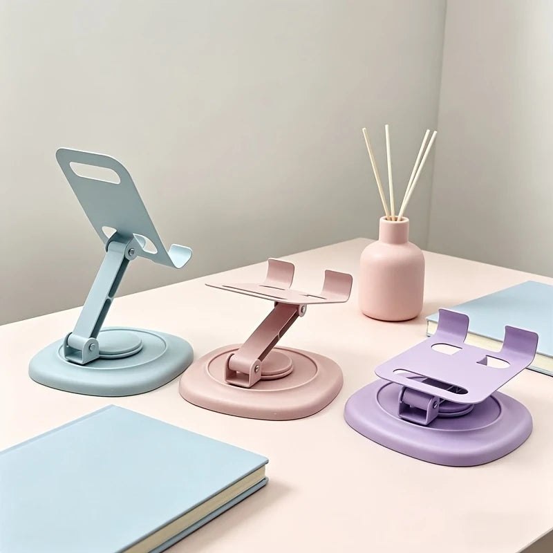A pink and blue 360° rotating foldable desktop phone stand is shown on a white table next to a notebook and chopsticks.